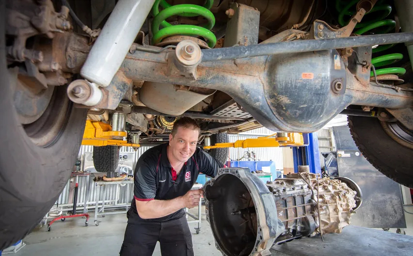 A mechanic lowering a gearbox from under a lifted 4WD with green coil springs